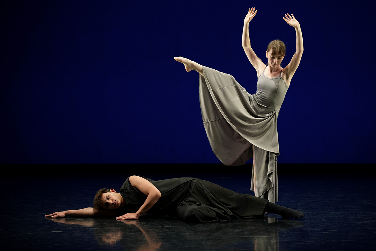 Two dancers on a stage with a dark blue backdrop: one lies on the floor in black while the other stands in a gray dress with one leg lifted high and both arms raised.