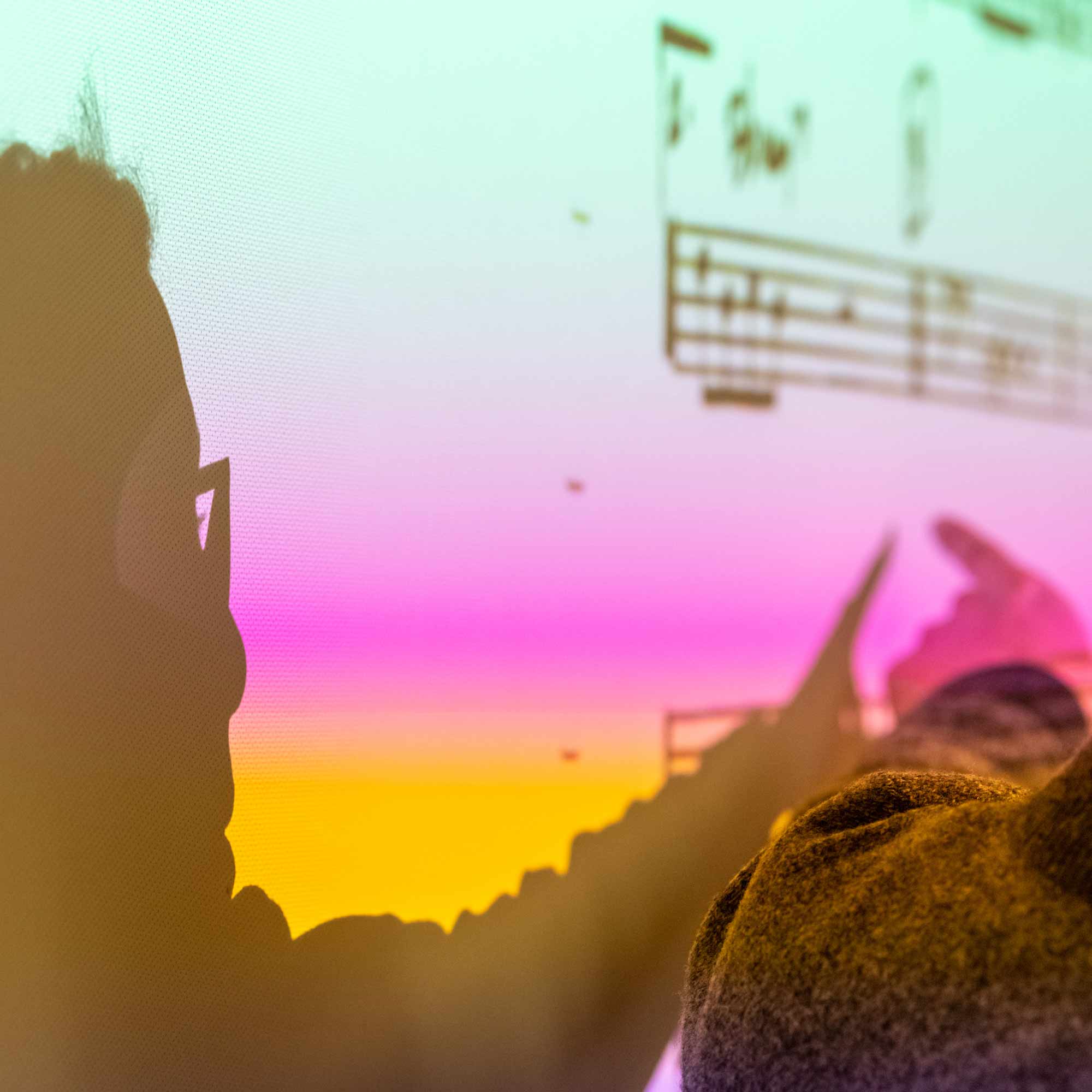  A person’s silhouette is visible in the foreground, facing right, wearing glasses, and pointing toward music notation on a large screen or monitor. The screen displays a vivid, pixelated gradient background of yellow, pink, and bright cyan.